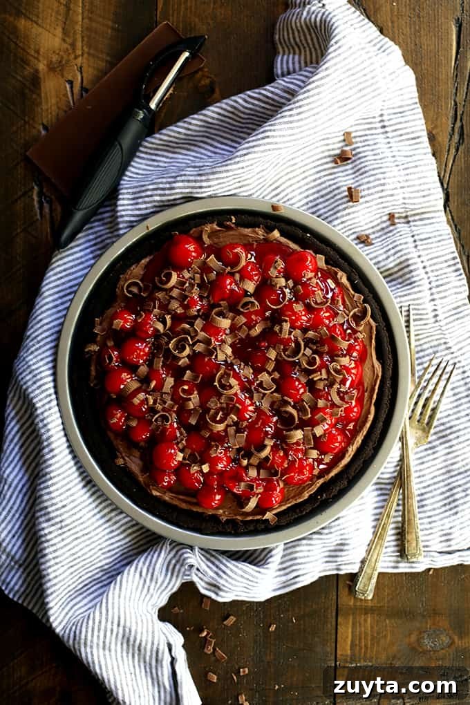 My Black Forest No Bake Cheesecake takes just 10 minutes to prepare and is an absolute must for chocolate lovers! A Black Forest No Bake Cheesecake sits on a striped napkin surrounded by two forks, a vegetable peeler and a bar of chocolate.