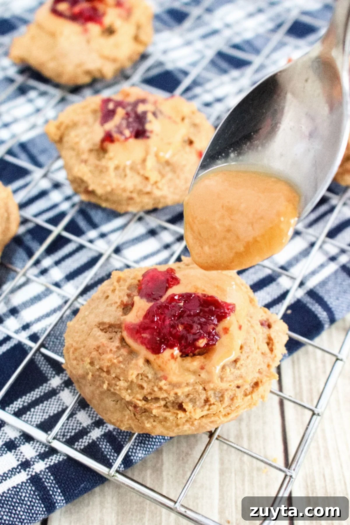 Flourless Peanut Butter and Jam Cookies (Dairy-Free Recipe) 2 Cooling rack on a blue linen with multiple peanut butter thumbprint cookies topped with jam and a spoon of peanut butter above the first.
