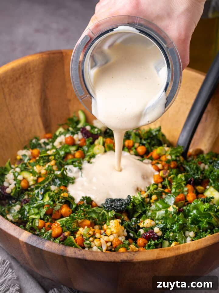 hand pouring cashew dressing on a kale and couscous salad with chickpeas and dried cranberries