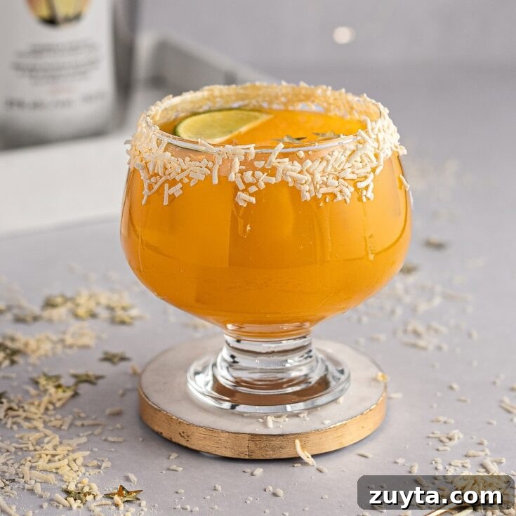 Mango tango drink on with a coconut glass rim, on a white and gold coaster, with gold stars and coconut scattered on the table.