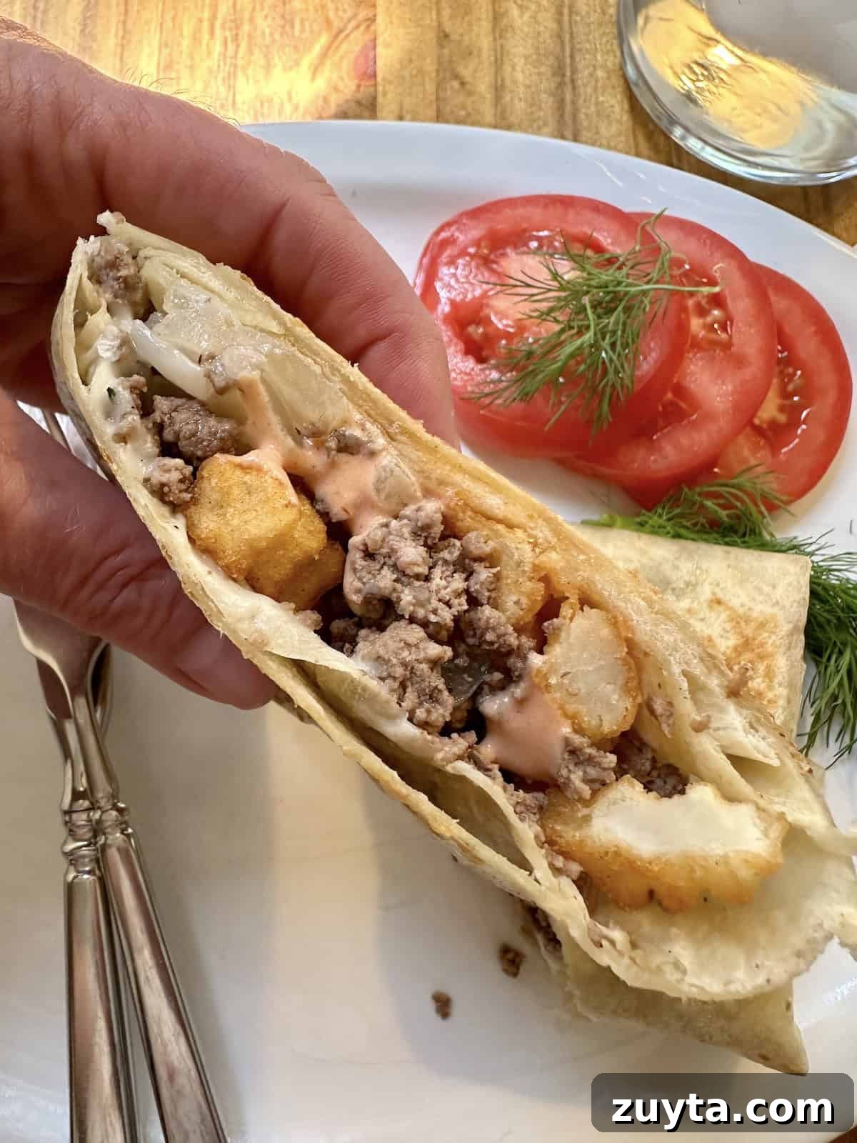 French Tacos Explained: Origins, Ingredients and Where to Try 2 One half of a French taco held by a hand over a plate with silverware, sliced tomato and dill.