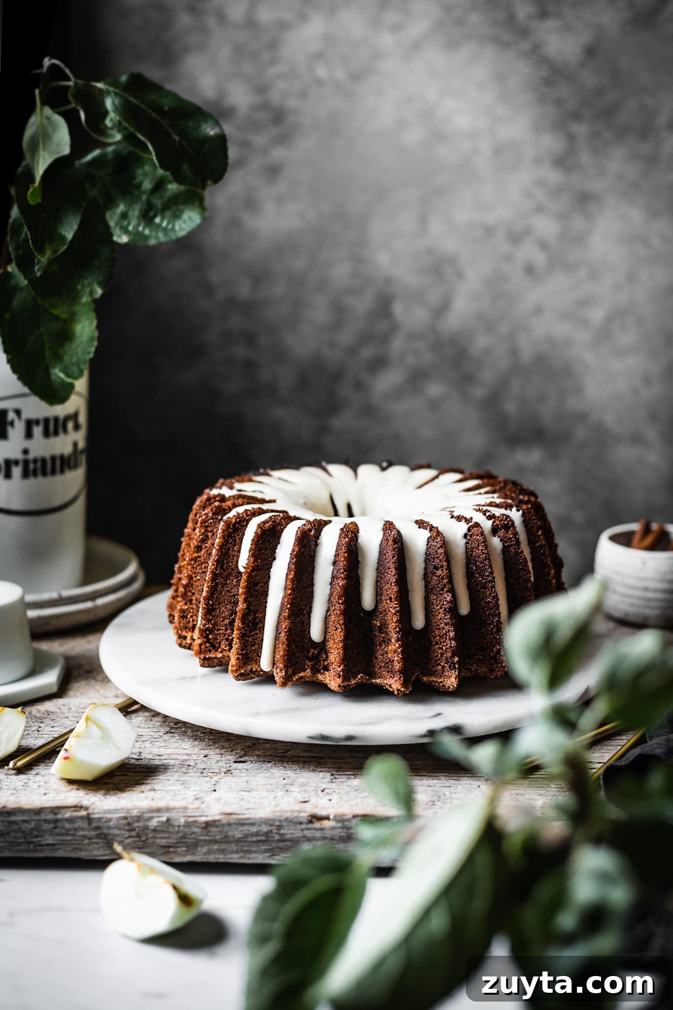 Moist Applesauce Bundt Cake Recipe with Cinnamon Glaze 5 Brown bundt cake with white glaze on a marble platter with apple branches in foreground
