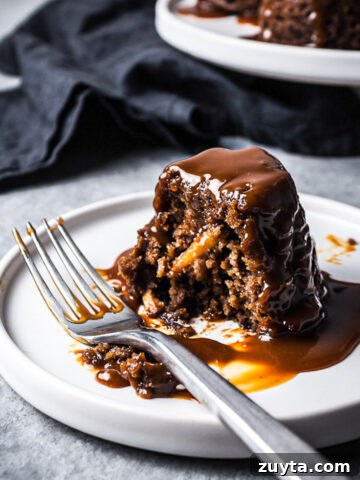 Close up of a mini cake on a white plate cut in half, revealing a moist crumb with bits of apples inside. The cake is covered in a drizzle of salted caramel sauce.