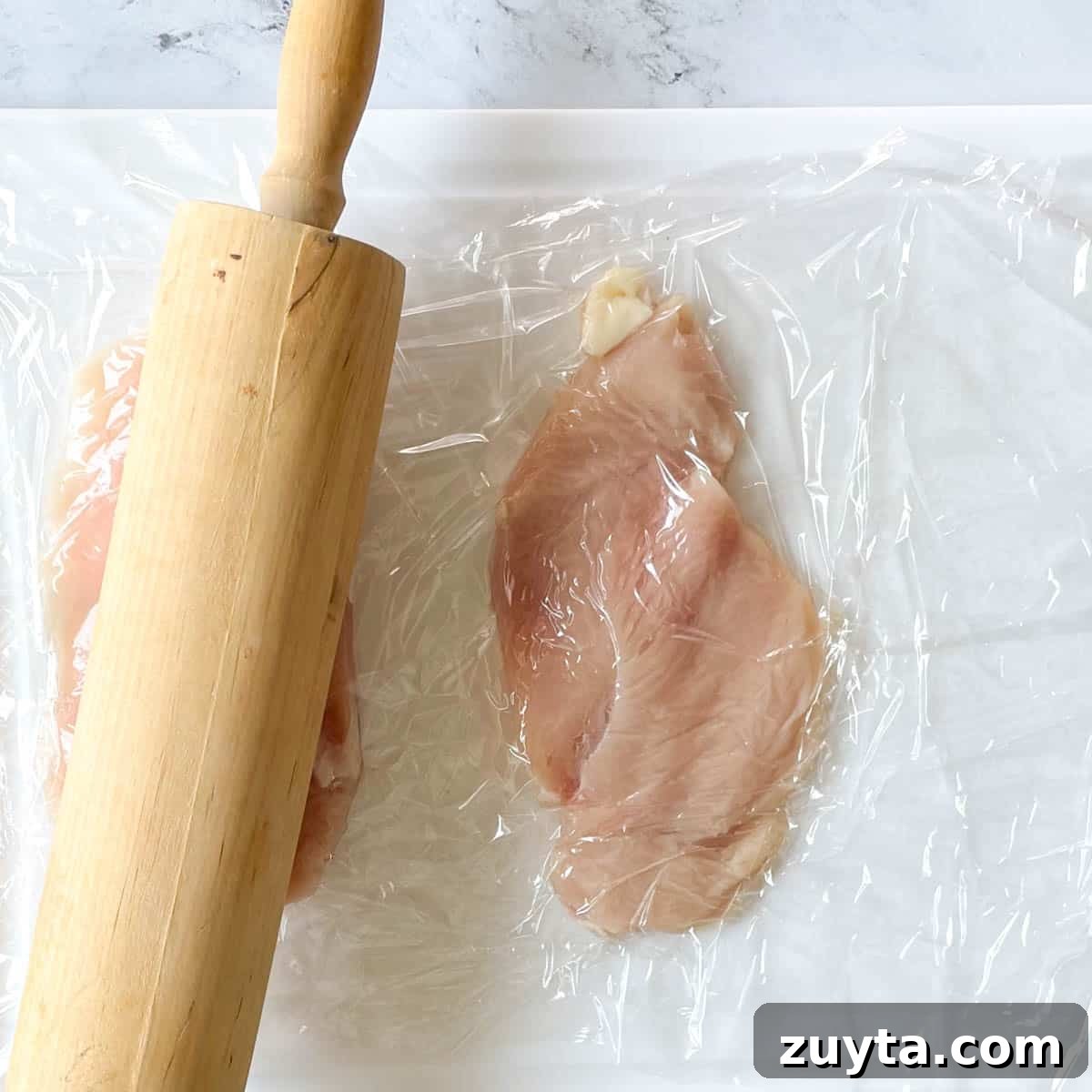 Crispy Italian Chicken Cutlets Recipe 4 Two cutlets are showing on a white cutting board covered with plastic wrap with a rolling pin flattening the cutlets.
