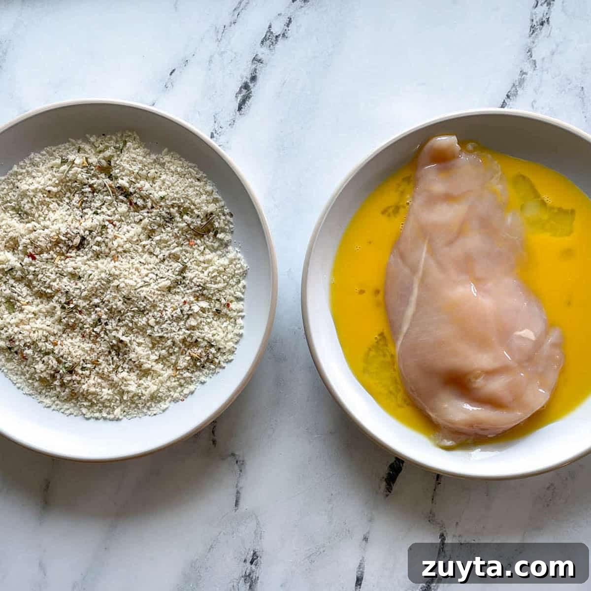 Crispy Italian Chicken Cutlets Recipe 5 One bowl with panko breadcrumbs and one bowl with beaten egg and a chicken breast on a marble counter.