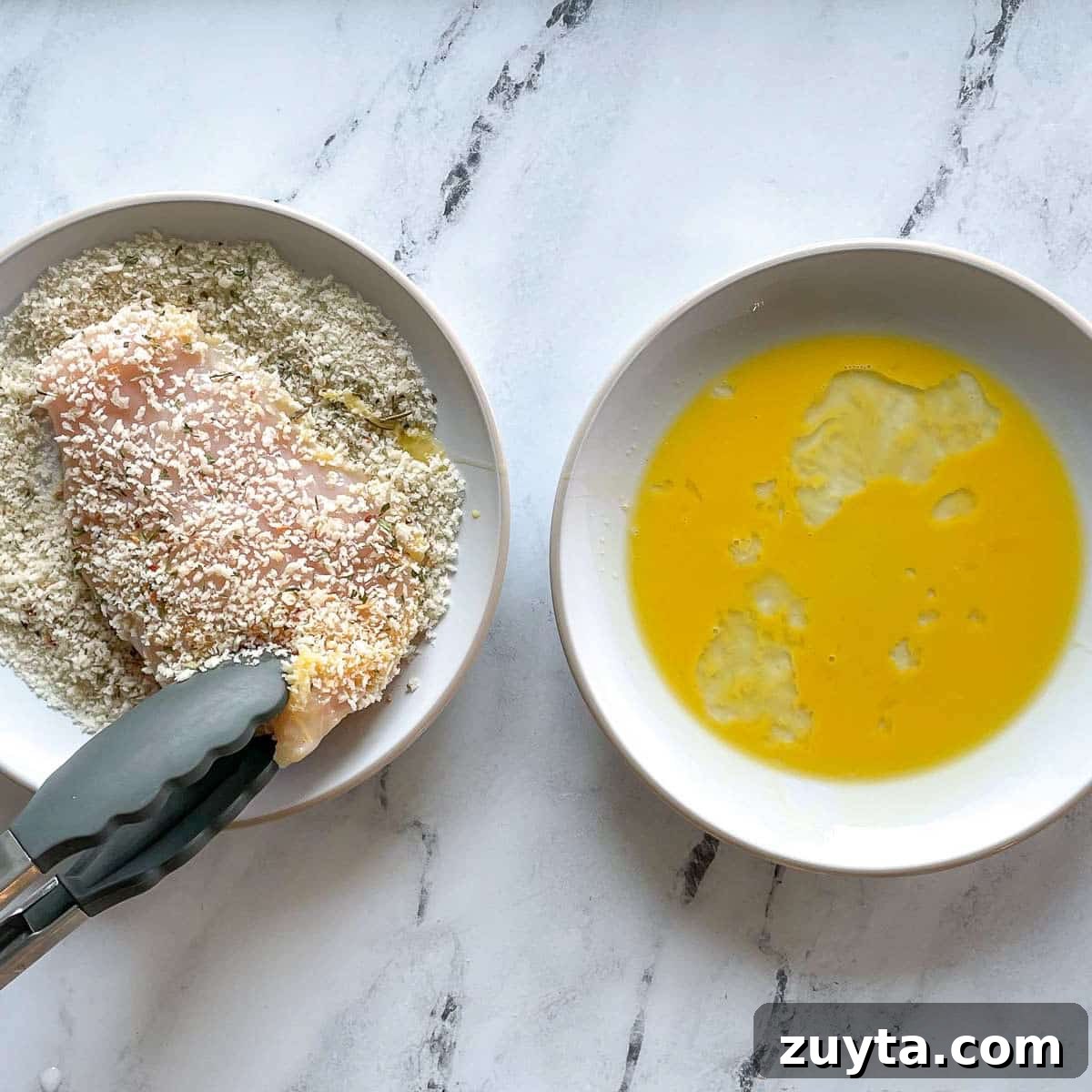 Crispy Italian Chicken Cutlets Recipe 6 One bowl with panko breadcrumbs and a breaded chicken cutlet and one bowl with beaten egg on a marble counter.