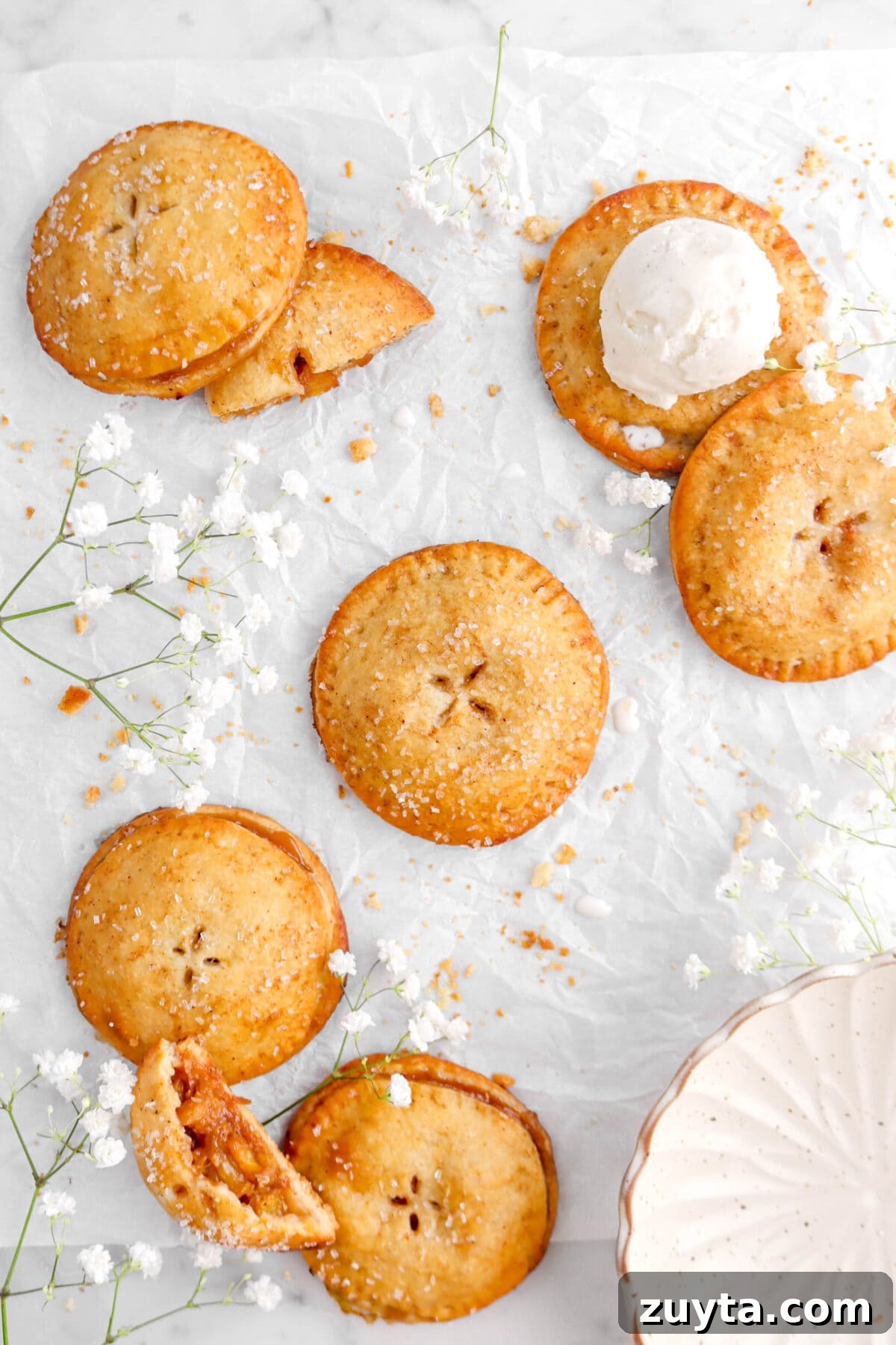 Cinnamon-Spiced Peach Hand Pies Recipe 13 pulled back shot of peach hand pies on parchment paper with one cut in half, white flowers around, a bowl, and scoop of ice cream on one hand pie