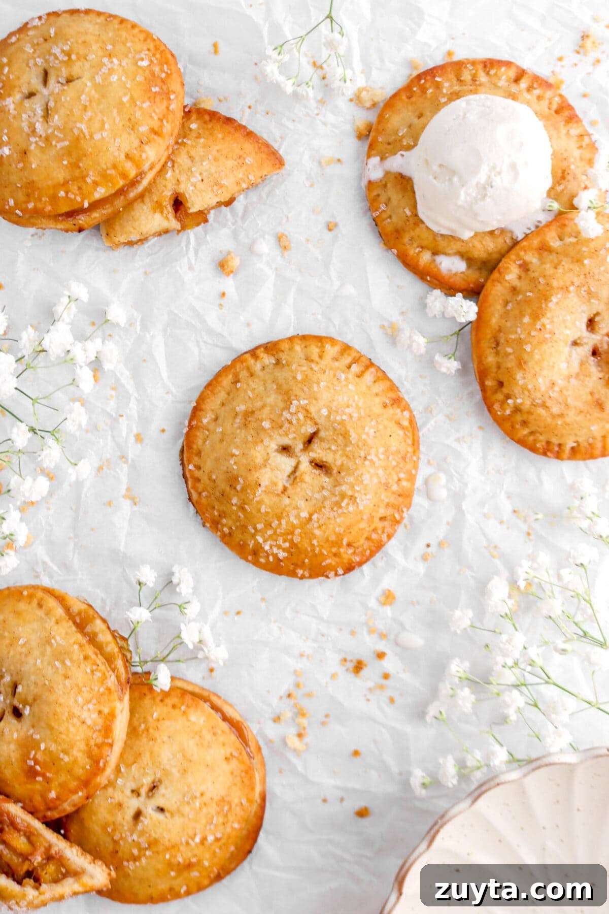 Cinnamon-Spiced Peach Hand Pies Recipe 3 pulled back shot of seven peach hand pies on crumbled parchment paper, with crumbs, white flowers, and a bowl around
