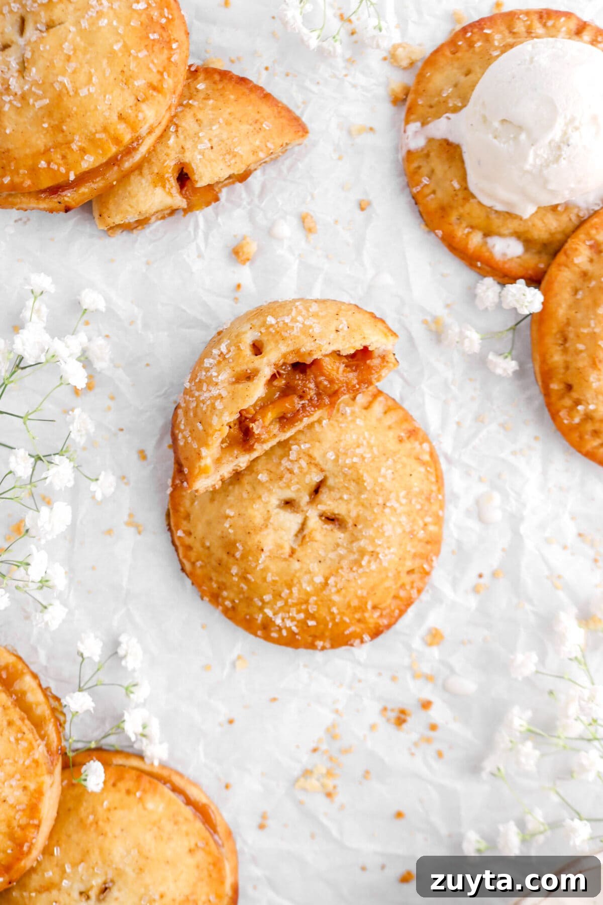 Cinnamon-Spiced Peach Hand Pies Recipe 10 hand pie on parchment paper with half leaning against it, white flowers, and more hand pies around on parchment paper