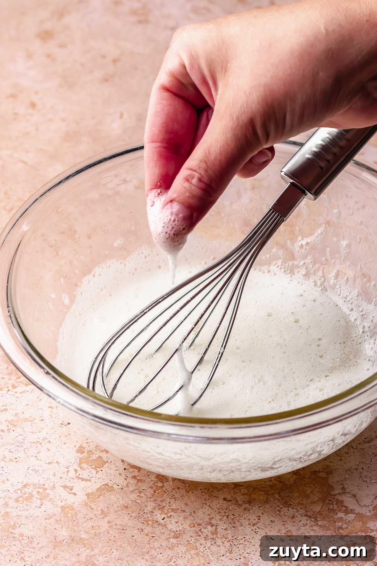 Silky Chocolate Swiss Meringue Buttercream Recipe 4 A hand dips fingers into a bowl of egg whites with a whisk sitting in it.