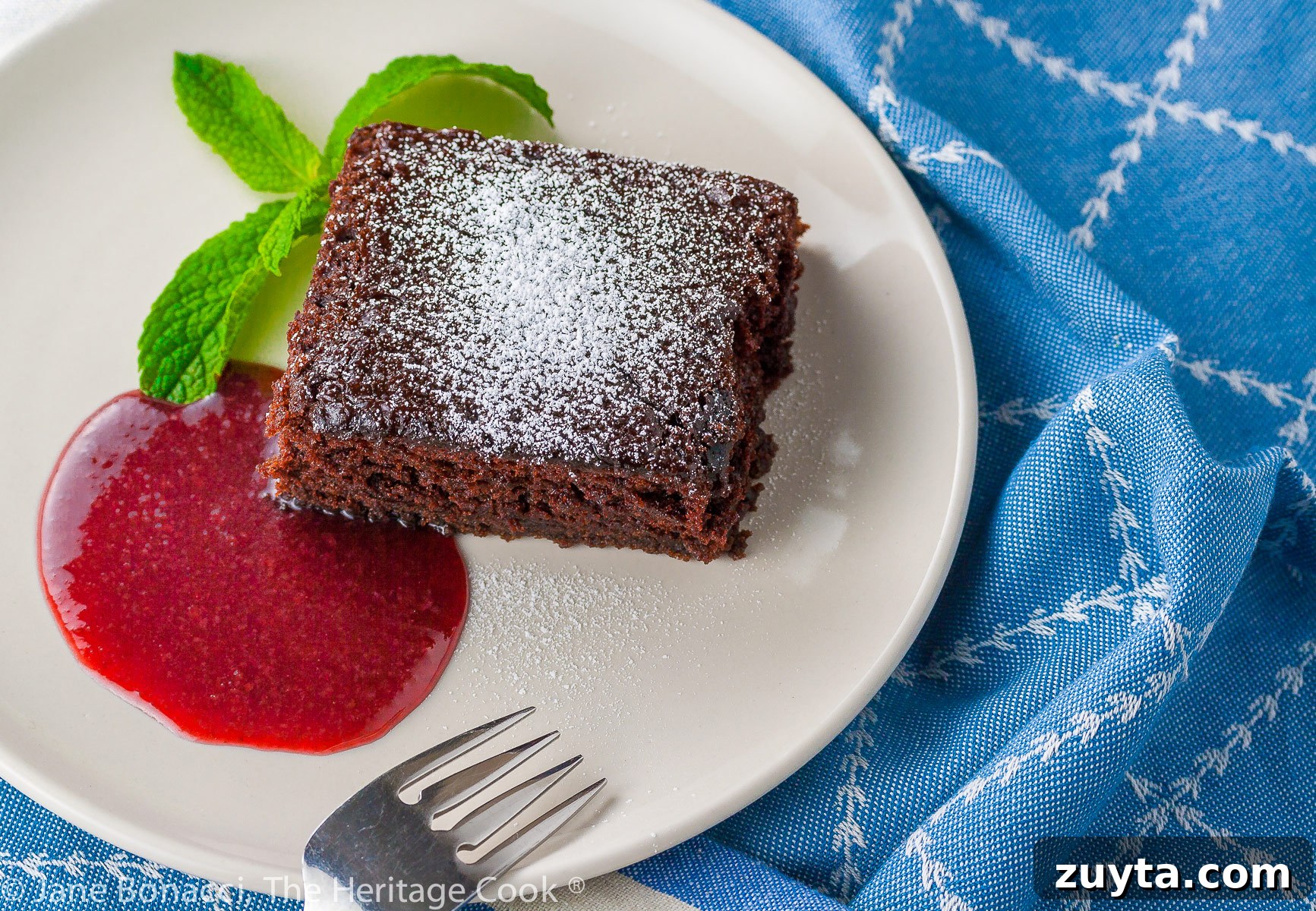 Gluten-Free Chocolate Cake with Raspberry Sauce 3 Square slice of chocolate cake surrounded by red raspberry sauce on a white plate with a mint sprig on blue background; Luscious Chocolate Cake with Raspberry Sauce © 2023 Jane Bonacci, The Heritage Cook.