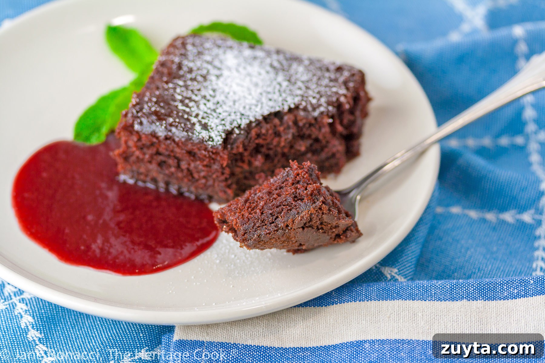Gluten-Free Chocolate Cake with Raspberry Sauce 4 Square slice of chocolate cake surrounded by red raspberry sauce on a white plate with a mint sprig on blue background; Luscious Chocolate Cake with Raspberry Sauce © 2023 Jane Bonacci, The Heritage Cook.