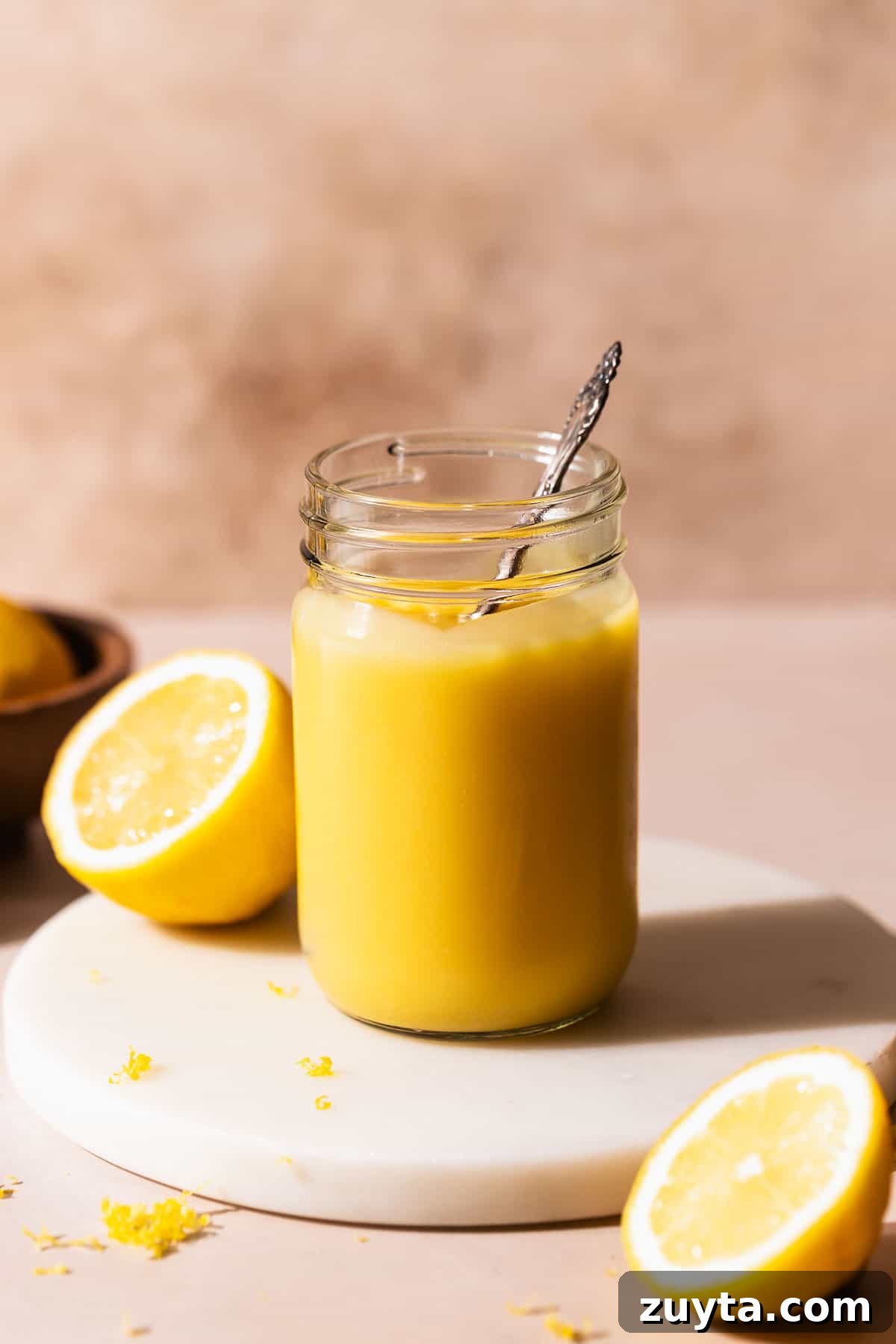 Lemon Curd Recipe: Homemade, Smooth, Zesty 2 A glass mason jar of homemade lemon curd on a white marble stand with a spoon dipping into it.