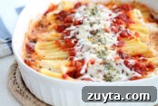 A white 9x13 oval baking dish filled with manicotti topped with red marinara sauce down the middle with mozzarella cheese and parsley on top sitting on a hot pad.