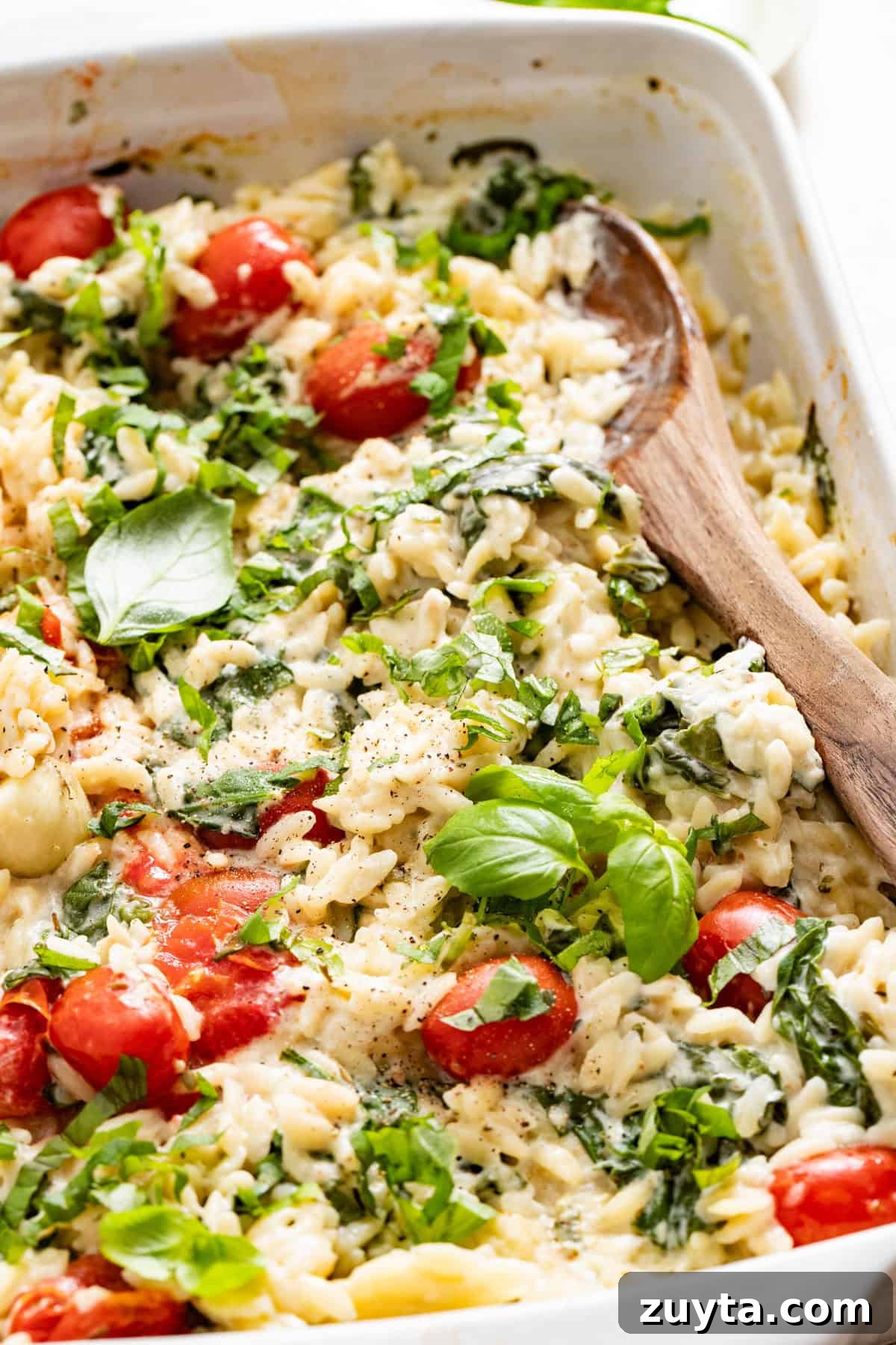 Creamy Boursin Orzo Bake Recipe 2 A dish filled with baked orzo topped with basil next to a wood spoon.
