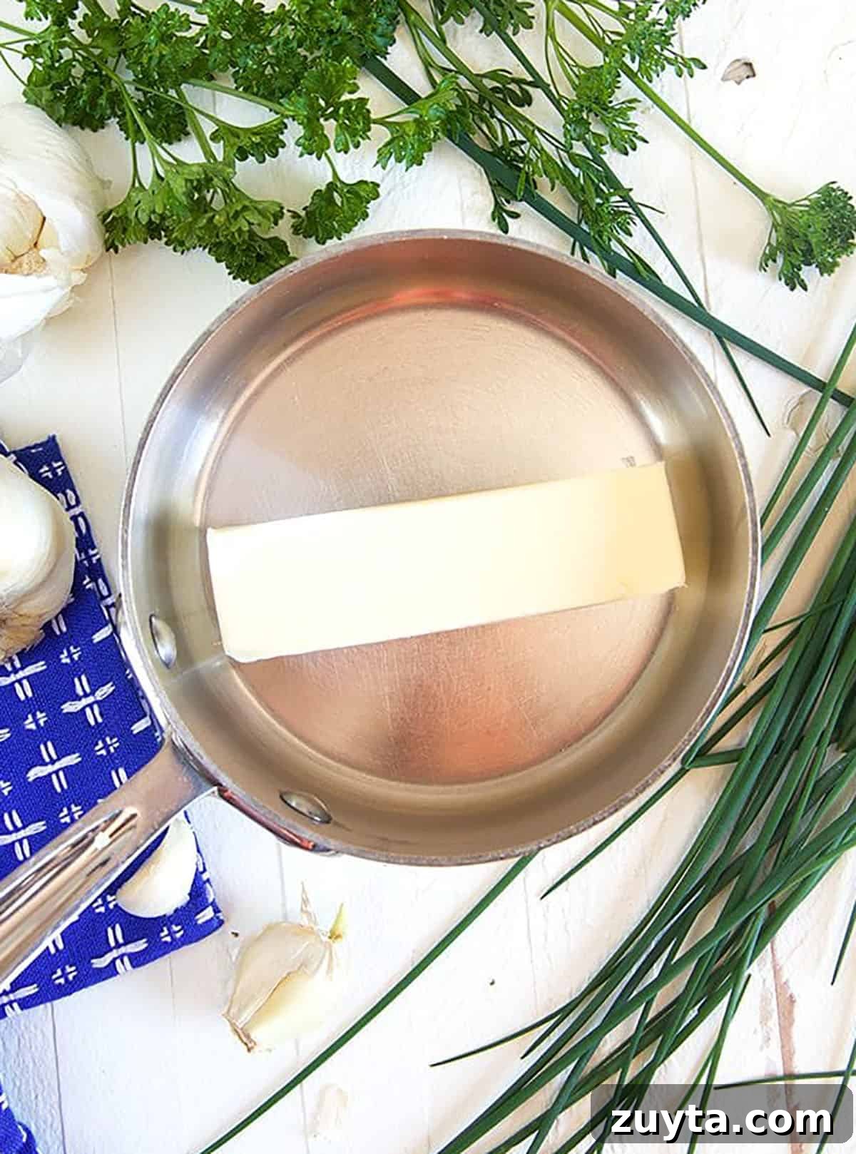 Stick of butter in a steel pot with herbs on a white background.