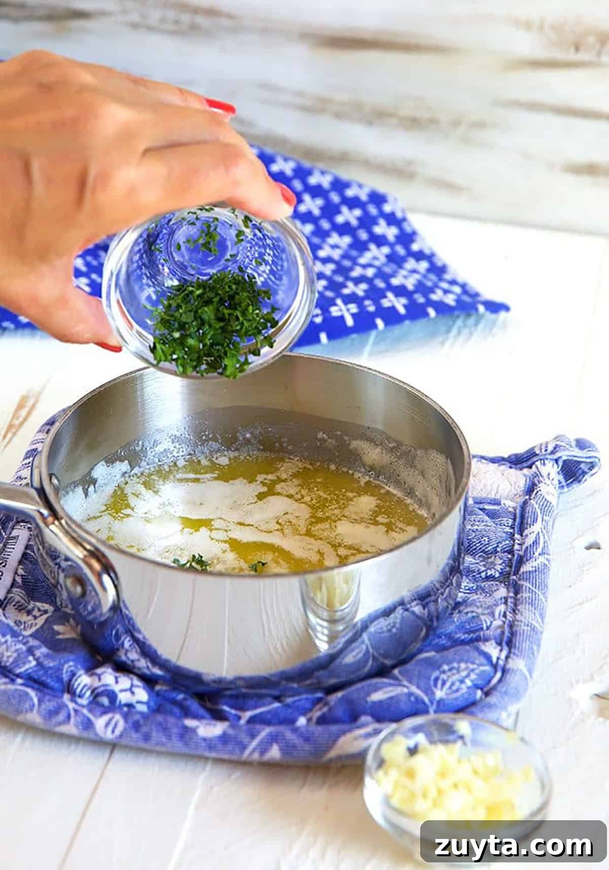 Melted butter in a skillet on a blue pot holder with herbs being poured into the butter.