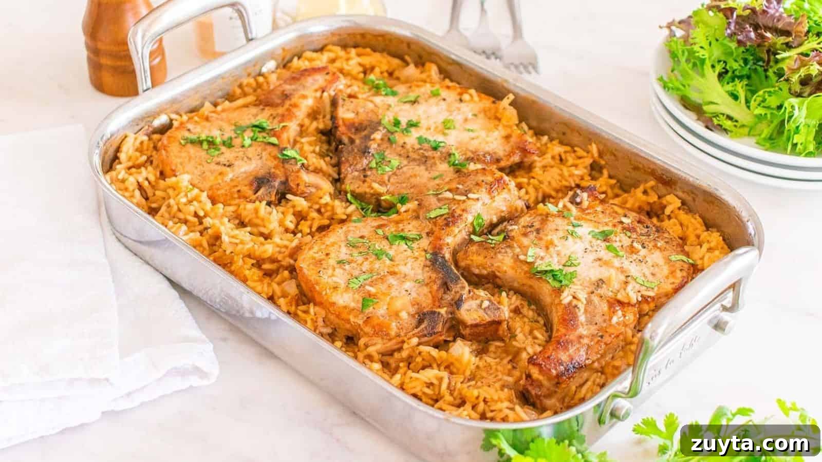 Foolproof Pork Chop Recipes for Juicy Weeknight Dinners 3 A baking dish with four cooked pork chops on top of seasoned rice, garnished with chopped herbs. Plates and a salad are in the background.