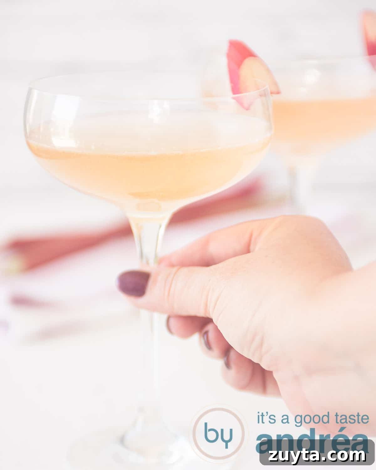 Sparkling Rhubarb Champagne Cocktail Recipe 5 An aerial view with two glasses of champagne rhubarb cocktail with a slice of rhubarb. A hand grabs the front glass.
