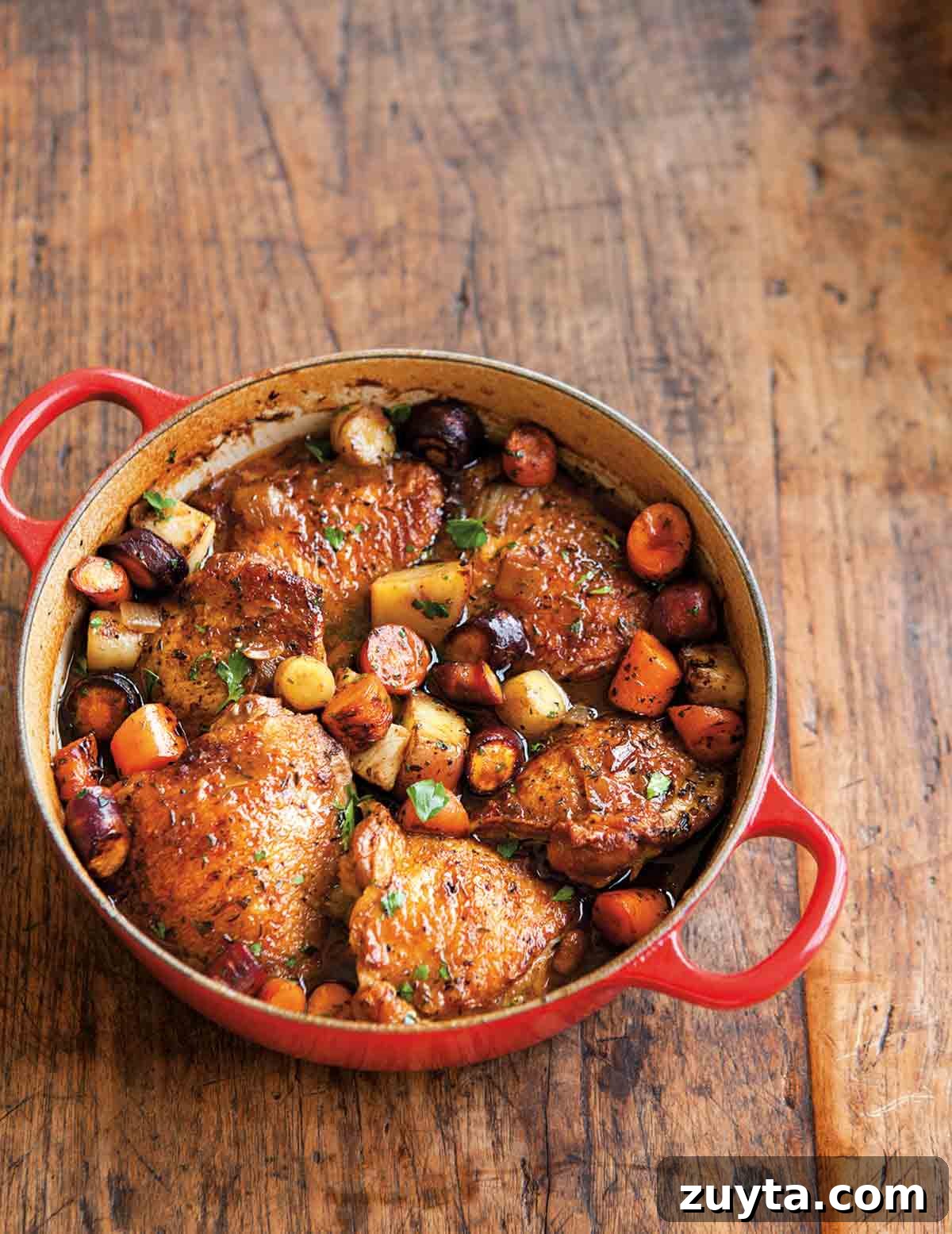 Braising Explained: What It Is and How to Braise 3 A red Dutch oven filled with beer-braised chicken with root vegetables on a wooden surface.