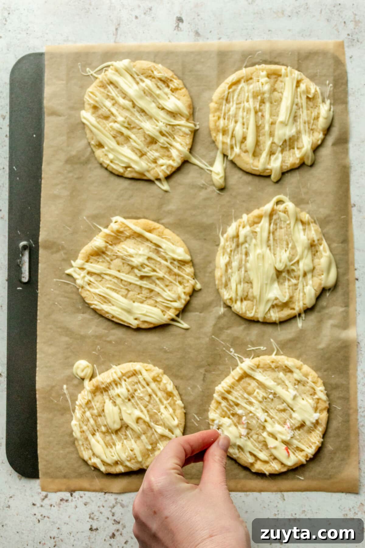 White Chocolate Peppermint Icebox Cookies Recipe 10 Crushed peppermints are tossed over white chocolate drizzled cookies on parchment paper on a baking tray on a light gray surface.