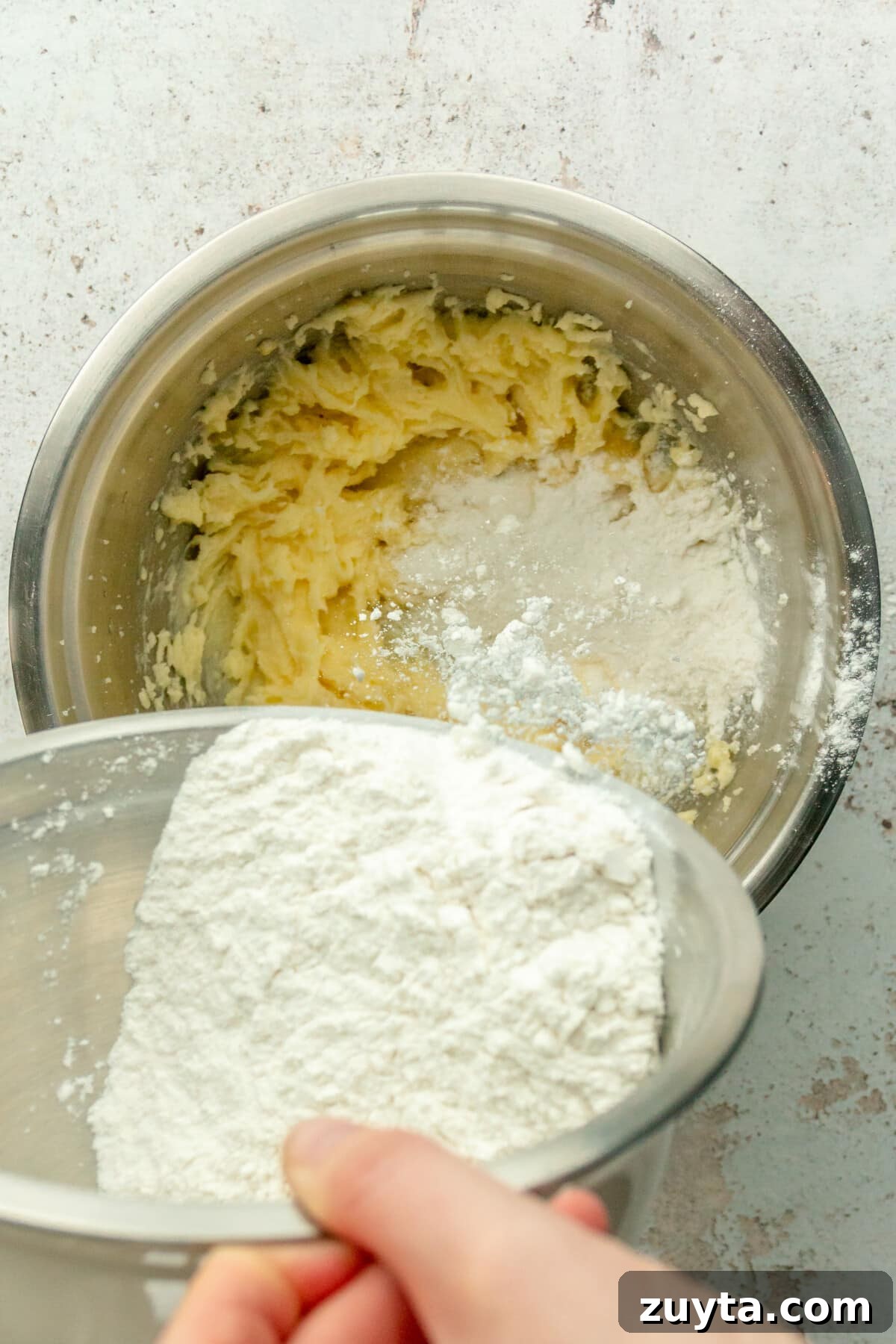 White Chocolate Peppermint Icebox Cookies Recipe 6 A mixture of flour and raising ingredients are tossed into a whipped butter mixture in a stainless steel bowl on a light gray surface.