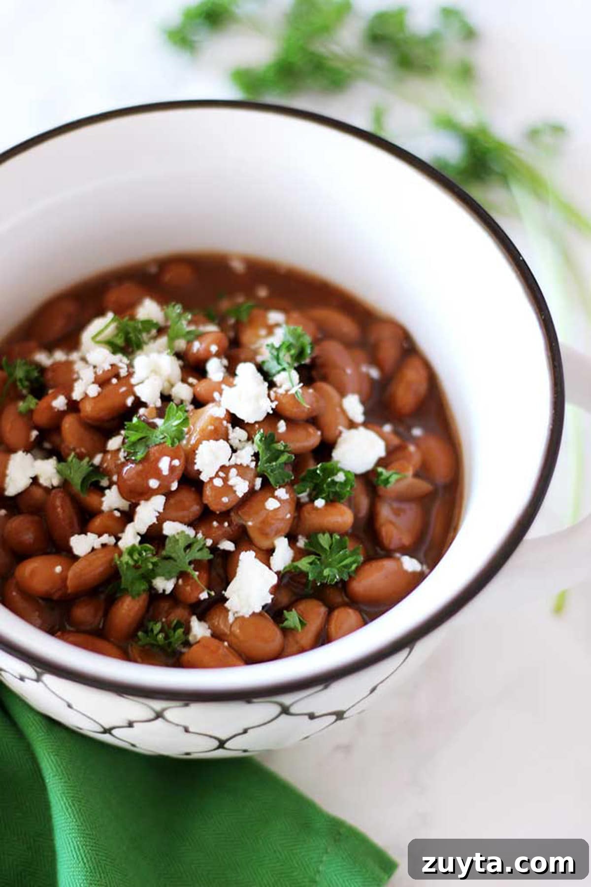 Slow Cooker Pinto Beans Recipe 2 A white bowl with pinto beans topped with crumbled feta cheese and parsley sitting on a white table with a green napkin.