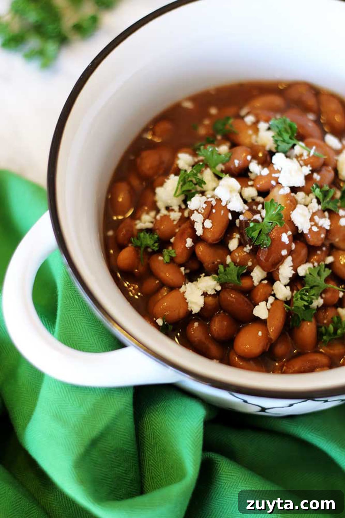 Slow Cooker Pinto Beans Recipe 3 A white bowl of pinto beans topped with crumbled feta cheese and parsley sitting on a green napkin.