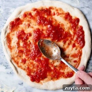 A close up of a pizza base getting tomato sauce spread on top before baking.