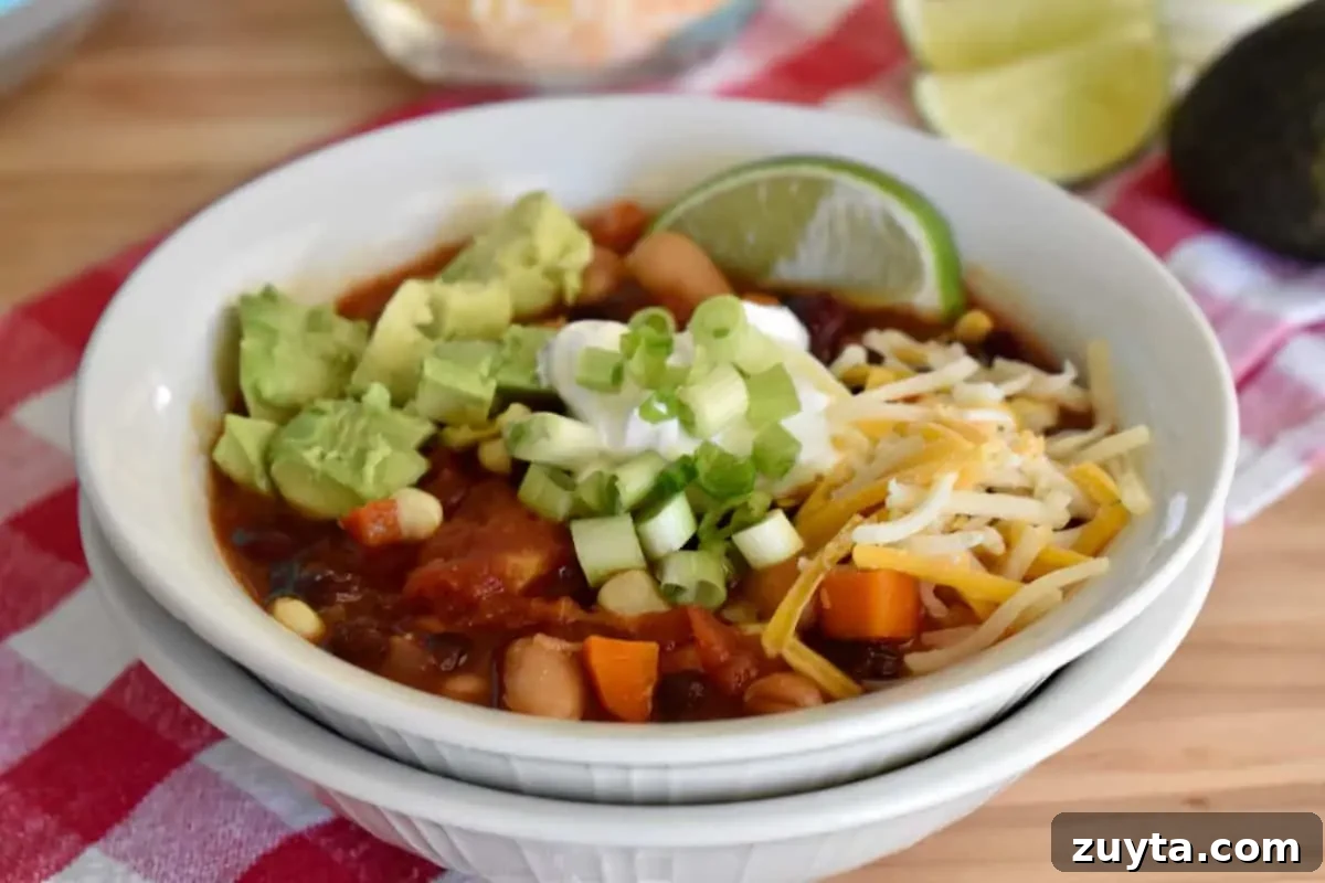 Slow Cooker Three-Bean Vegetarian Chili Recipe 6 White bowl filled with three bean vegetarian chili topped with green onion, avocado, cheese, and a lime wedge.