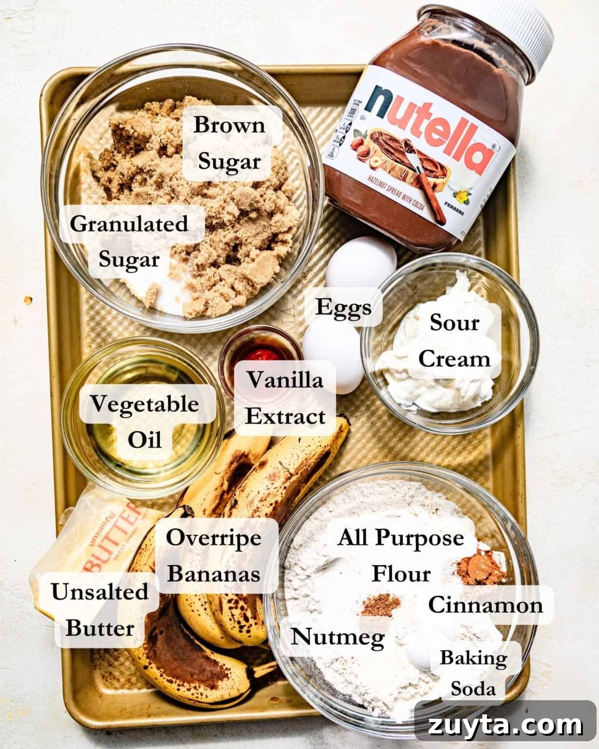 ingredients to make banana nutella muffins on a baking sheet - nutella, overripe bananas, flour, kosher salt, cinnamon, nutmeg, sugar, brown sugar, eggs, sour cream, oil, and vanilla extract.