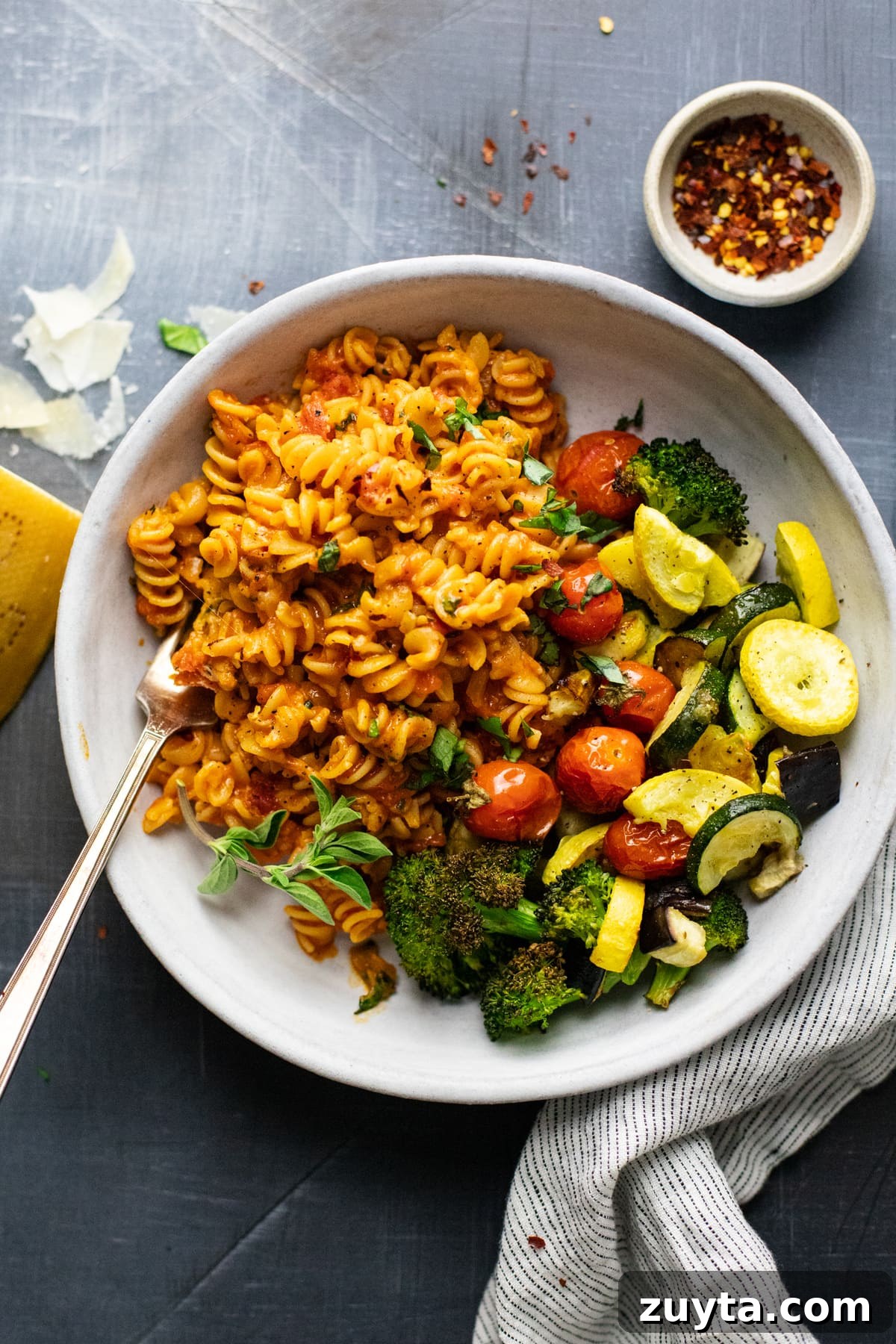 white bowl with rotini pasta noodles and roasted vegetables in it on gray background with shredded cheese and red pepper flakes arranged around it.