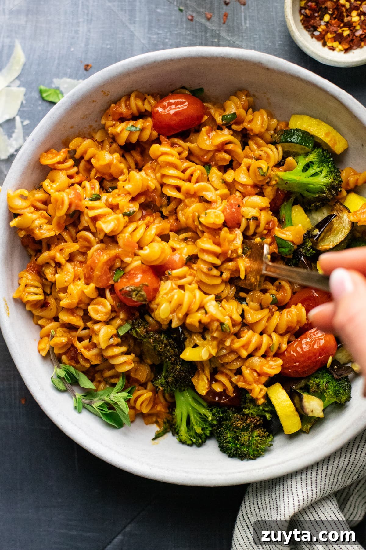 hand holding fork in white bowl of rotini pasta noodles and roasted vegetables.