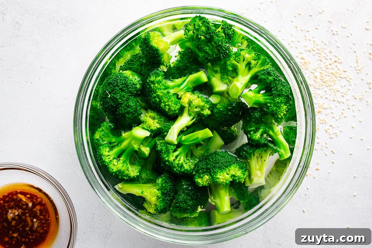 Blanched broccoli florets in an ice bath next to a smaller bowl with sesame dressing, viewed from above.