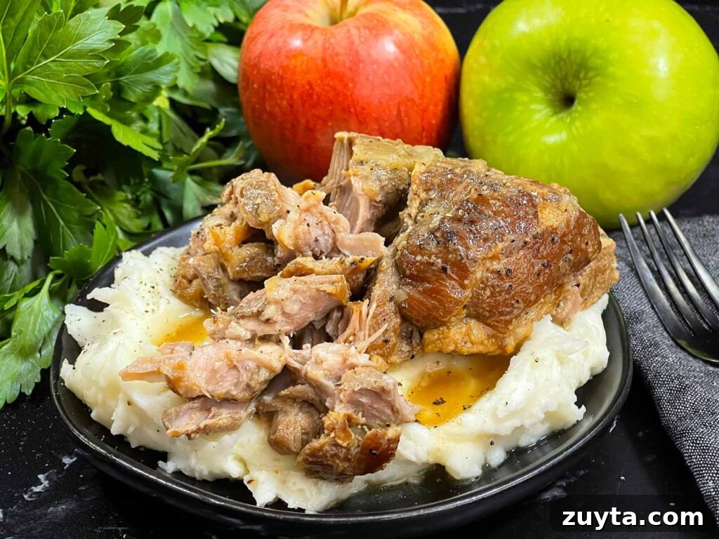 Slow-Braised Apple Cider Pork Shoulder 9 A plate of apple cider braised pork shoulder on a bed of mashed potatoes on a dark plate. Parsley and apples are in the background.