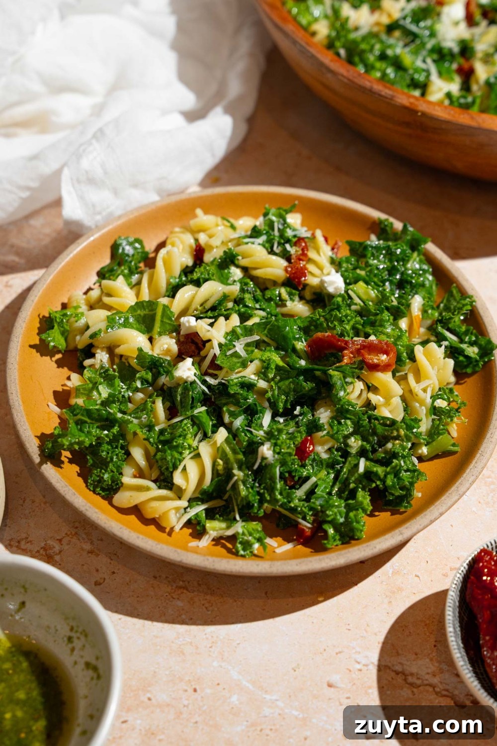 Sam's Club Copycat Mediterranean Pasta Salad Recipe 2 A plate with Mediterranean pasta salad garnished with grated parmesan. The salad consists of kale, rotini pasta, and sundried tomatoes. The plate is on a tan counter with a wooden bowl of pasta salad and white cloth napkin.