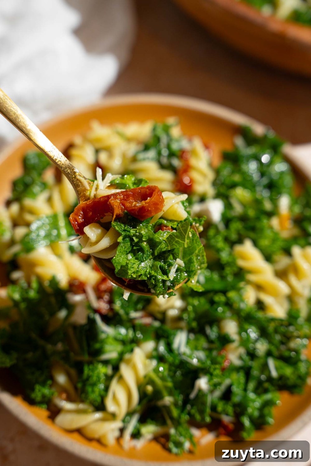 Sam's Club Copycat Mediterranean Pasta Salad Recipe 11 A close up of a fork with a bite of Mediterranean pasta salad. There is a wooden bowl of pasta salad on a tan counter with a white cloth napkin blurred in the background.