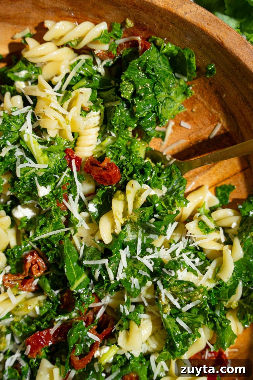 Sam's Club Copycat Mediterranean Pasta Salad Recipe 3 Mediterranean pasta salad served in a wooden bowl garnished with grated parmesan. The salad includes kale, rotini pasta, and sundried tomatoes. There is a fork in the bowl.