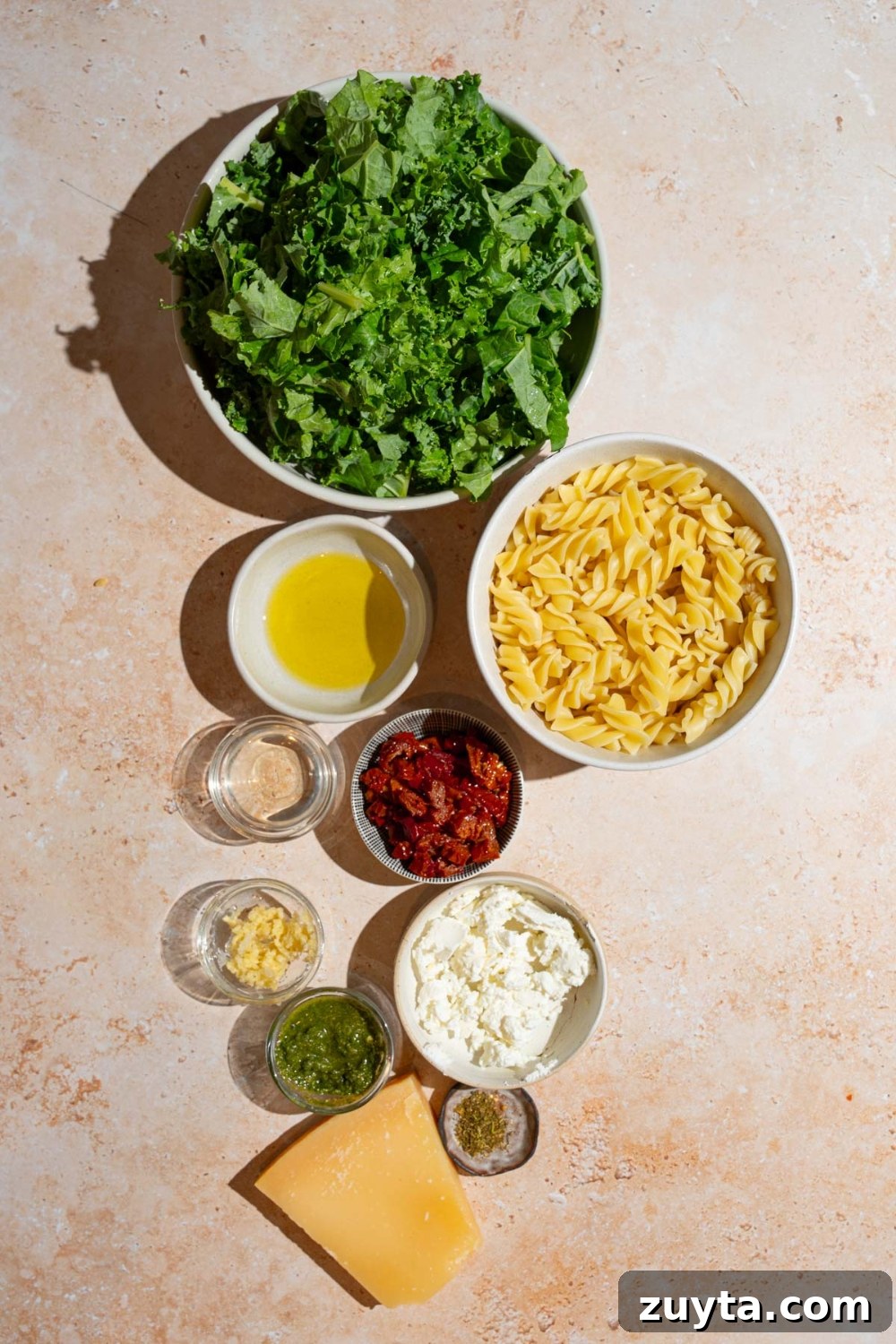 Sam's Club Copycat Mediterranean Pasta Salad Recipe 4 An overhead shot of several bowls in various sizes containing ingredients to make Mediterranean pasta salad including chopped kale, rotini pasta, sundried tomatoes, olive oil, pesto, balsamic vinegar, garlic, feta cheese, and seasonings.