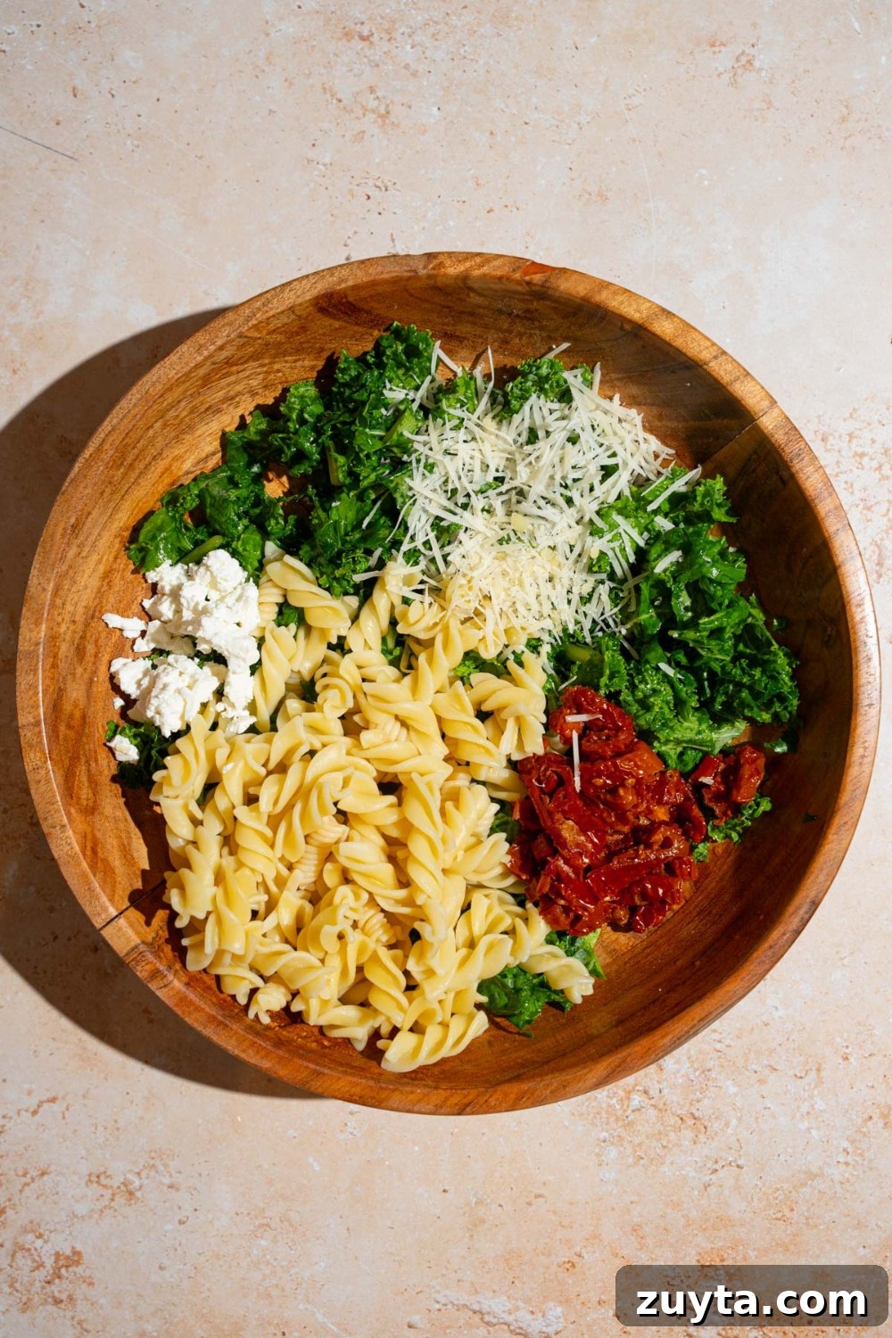 Sam's Club Copycat Mediterranean Pasta Salad Recipe 7 A wooden bowl with ingredients to make Mediterranean pasta salad including kale, rotini pasta, sundried tomatoes, feta cheese, and grated parmesan. The bowl is on a tan counter.