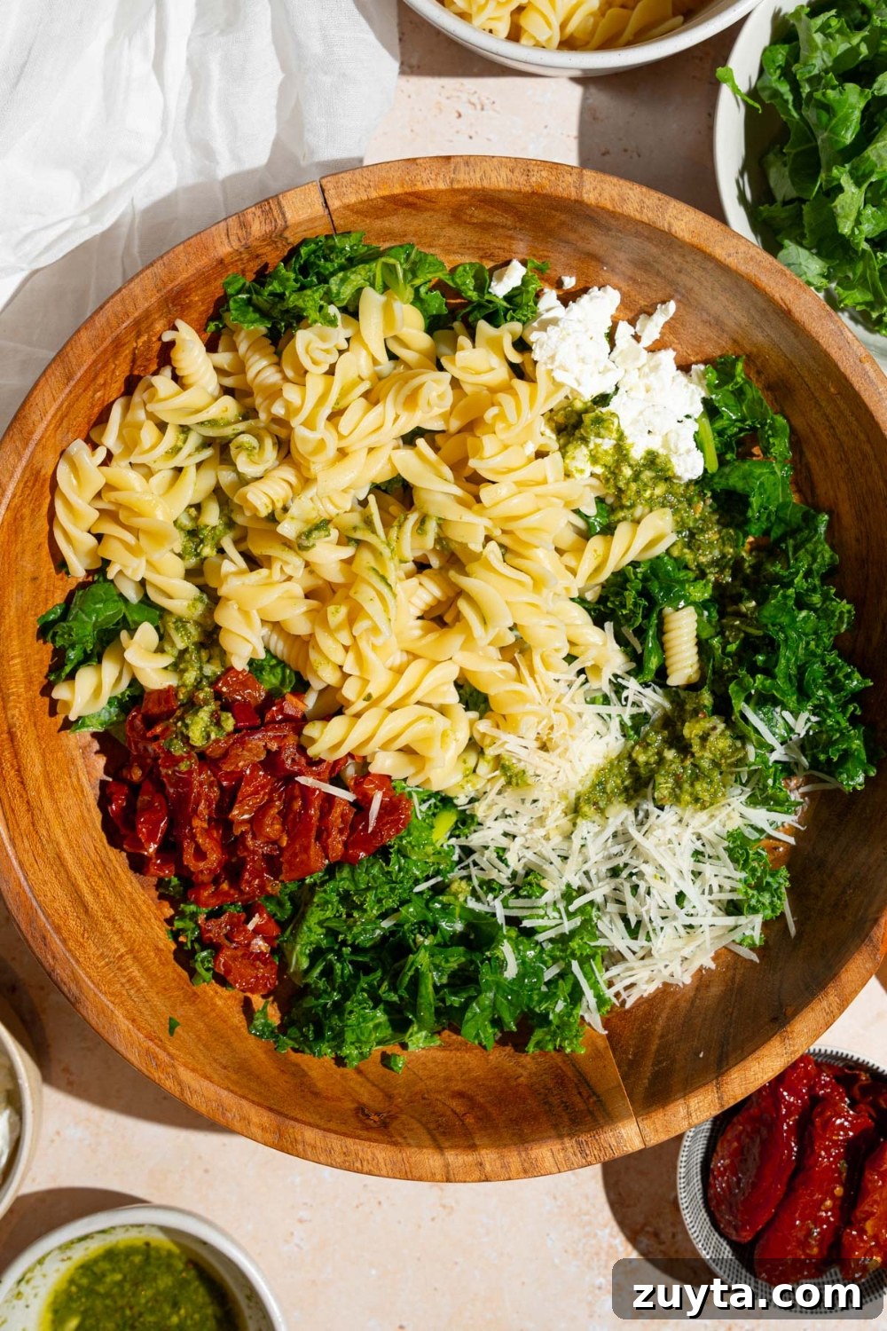 Sam's Club Copycat Mediterranean Pasta Salad Recipe 8 A wooden bowl with ingredients to make Mediterranean pasta salad including kale, rotini pasta, sundried tomatoes, feta cheese, and grated parmesan. The bowl is on a tan counter with a white cloth napkin and bowls with salad ingredients.