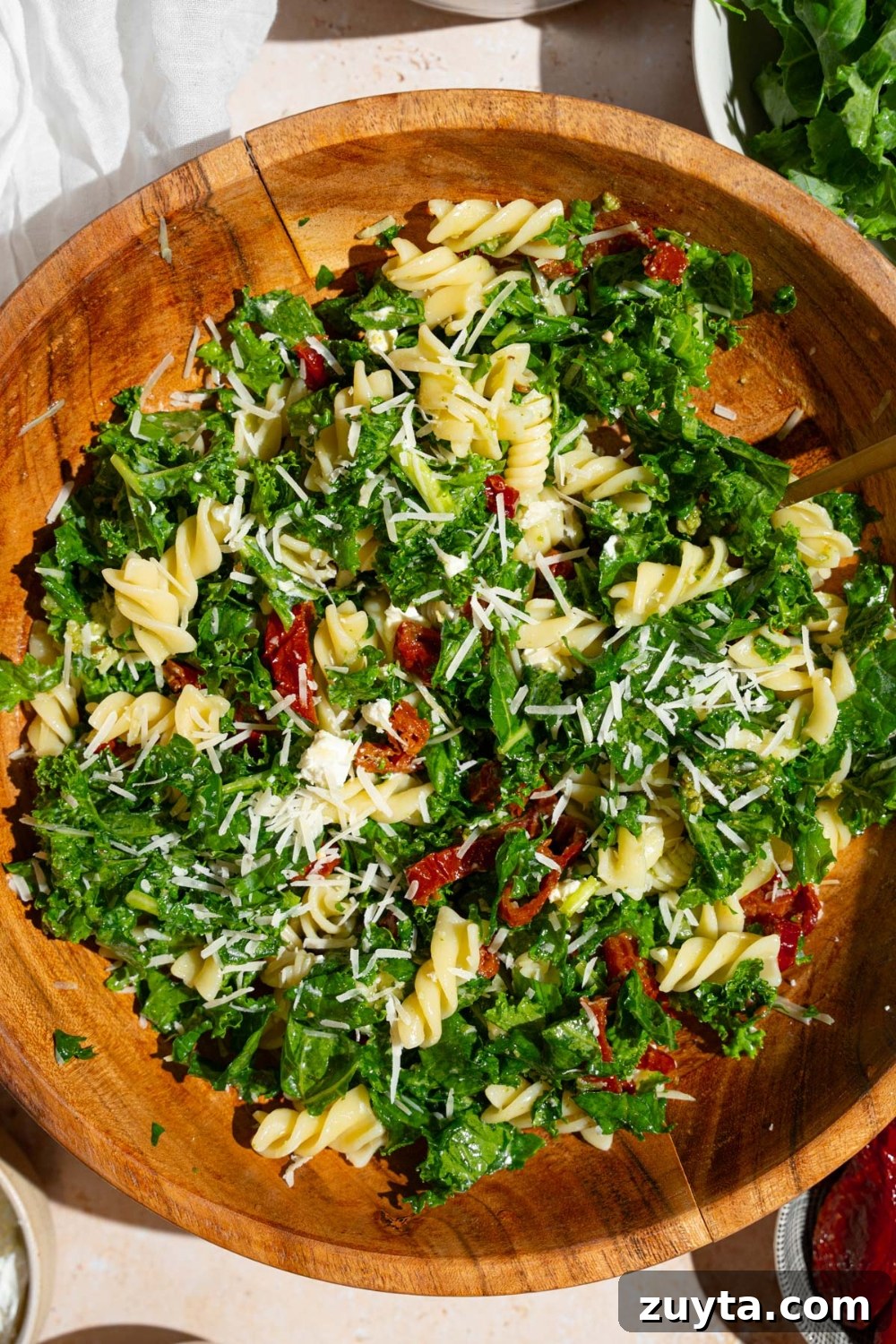 Sam's Club Copycat Mediterranean Pasta Salad Recipe 9 Mediterranean pasta salad served in a wooden bowl garnished with grated parmesan. The salad includes kale, rotini pasta, and sundried tomatoes. There is a fork in the bowl.