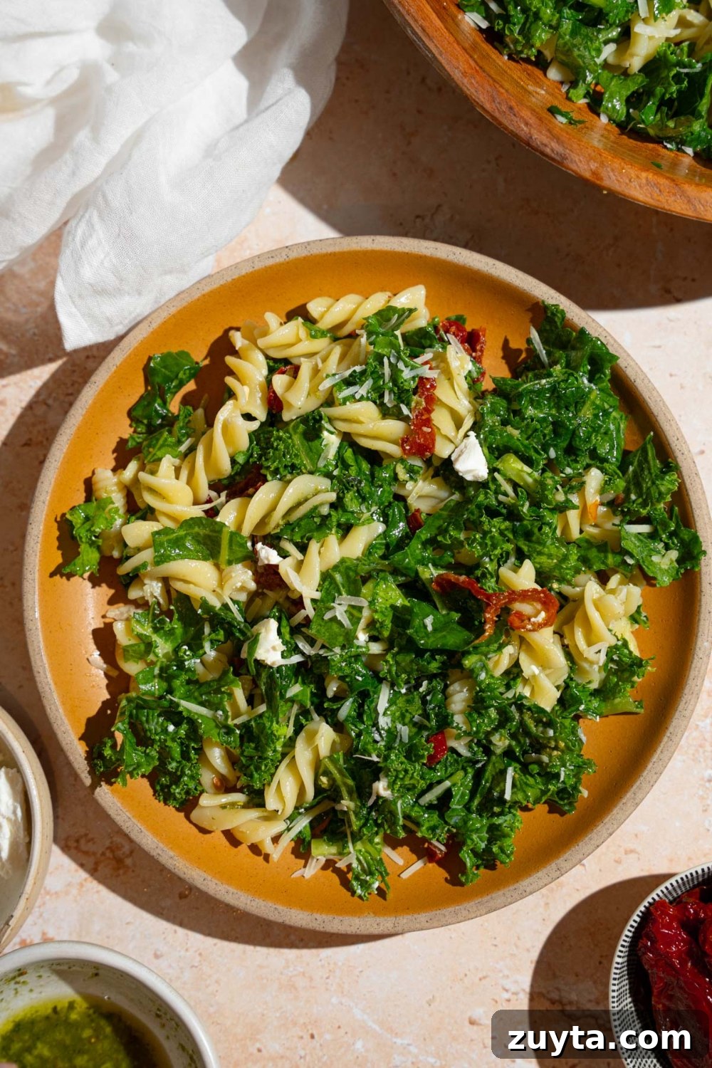 Sam's Club Copycat Mediterranean Pasta Salad Recipe 10 A plate with Mediterranean pasta salad garnished with grated parmesan. The salad consists of kale, rotini pasta, and sundried tomatoes. The plate is on a tan counter with a wooden bowl of pasta salad and white cloth napkin.