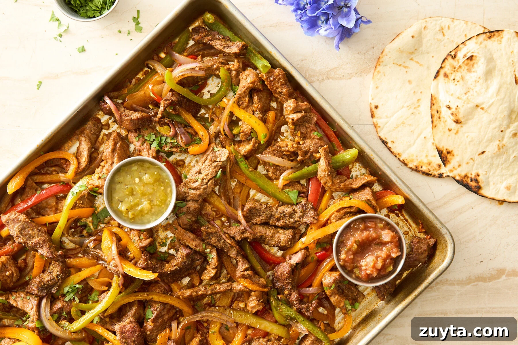 Sheet pan steak fajitas on a baking sheet with salsa verde and red salsa next to two tortillas. 