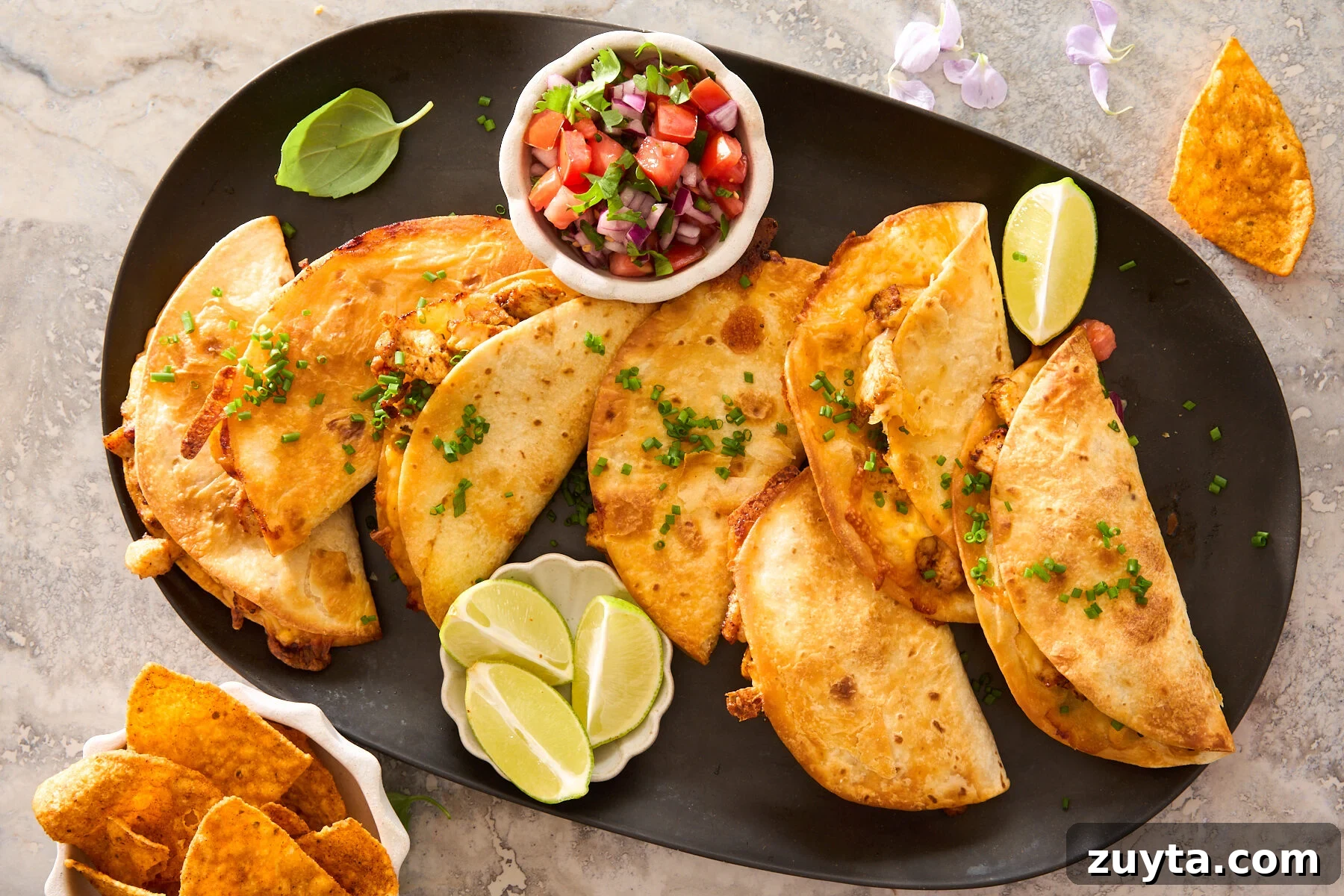 A black platter with six crispy tacos garnished with chopped chives, served with lime wedges and a small bowl of fresh tomato salsa. Tortilla chips sit in a dish nearby.