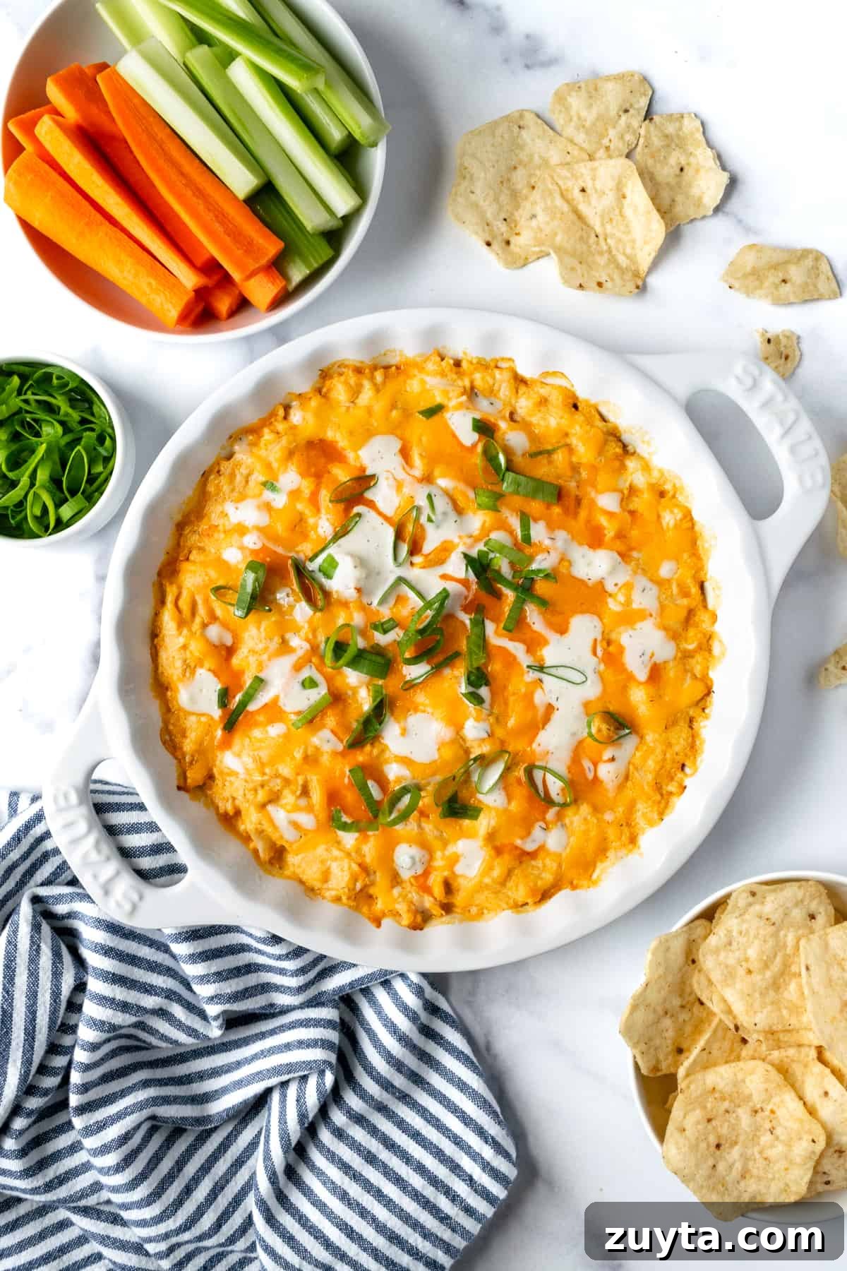 Creamy Buffalo Chicken Dip with Greek Yogurt 2 Greek Yogurt Buffalo Chicken Dip in a White Dish on a countertop with chips and veggies for dipping.