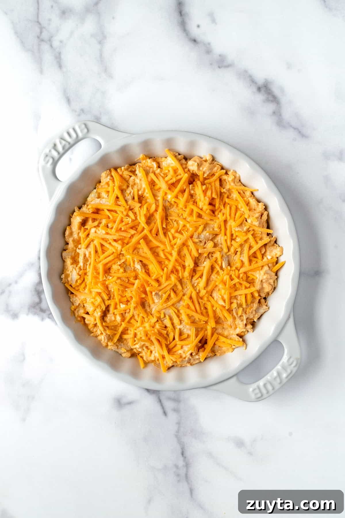 Creamy Buffalo Chicken Dip with Greek Yogurt 6 Buffalo Chicken Dip with Greek Yogurt Topped with Cheddar Cheese in a White Ceramic Baking Dish Ready to be baked.