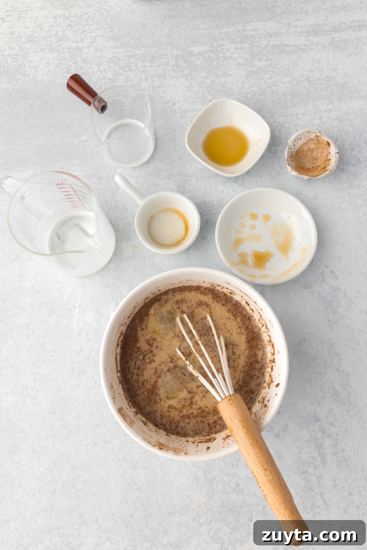 Cinnamon Roll Casserole Recipe with Cream Cheese Glaze 7 Whisking together ingredients in a bowl, with empty cups and bowls surrounding it.