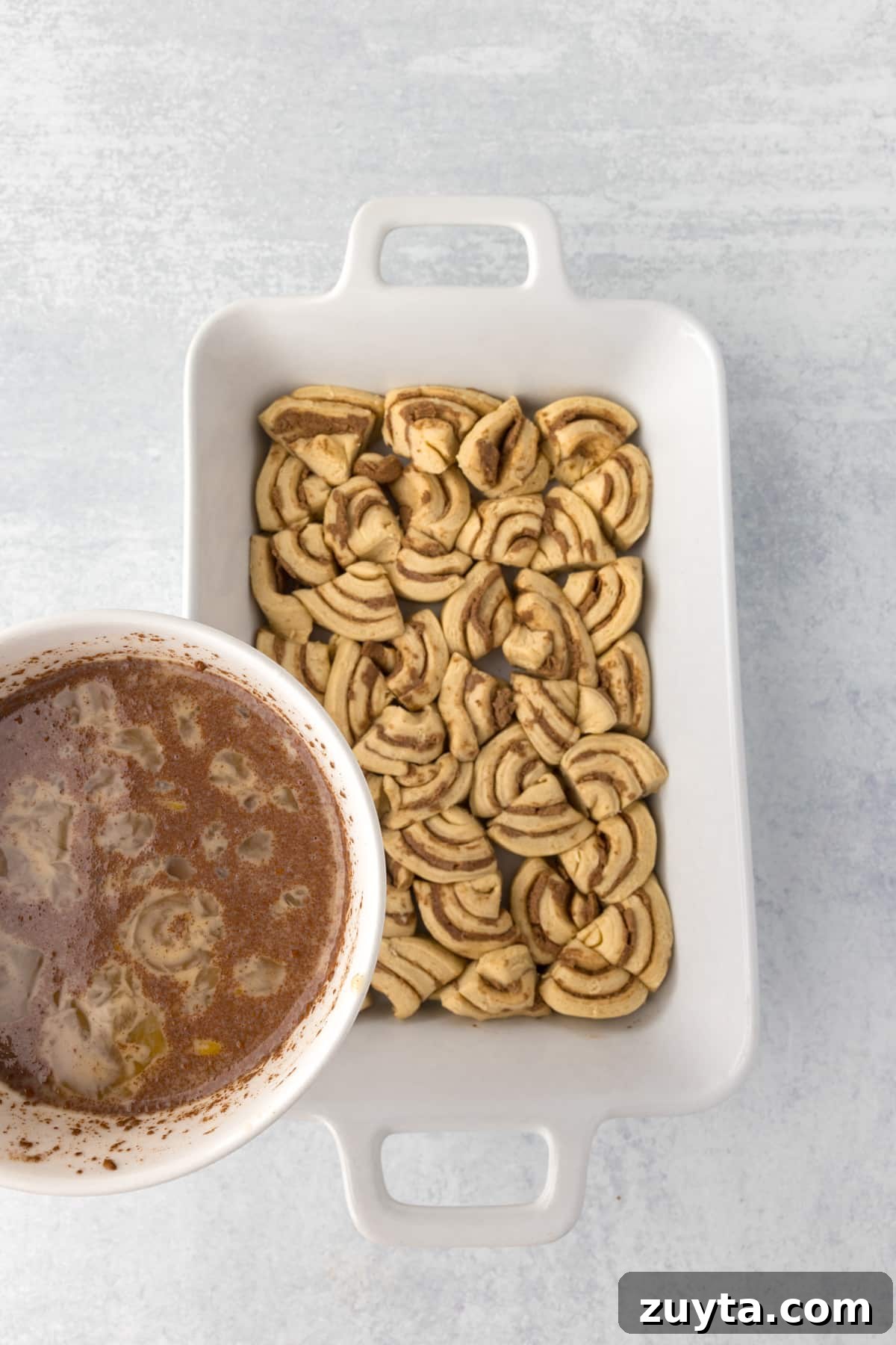 Cinnamon Roll Casserole Recipe with Cream Cheese Glaze 8 Pouring a custard mixture over a baking dish filled with cinnamon roll pieces.