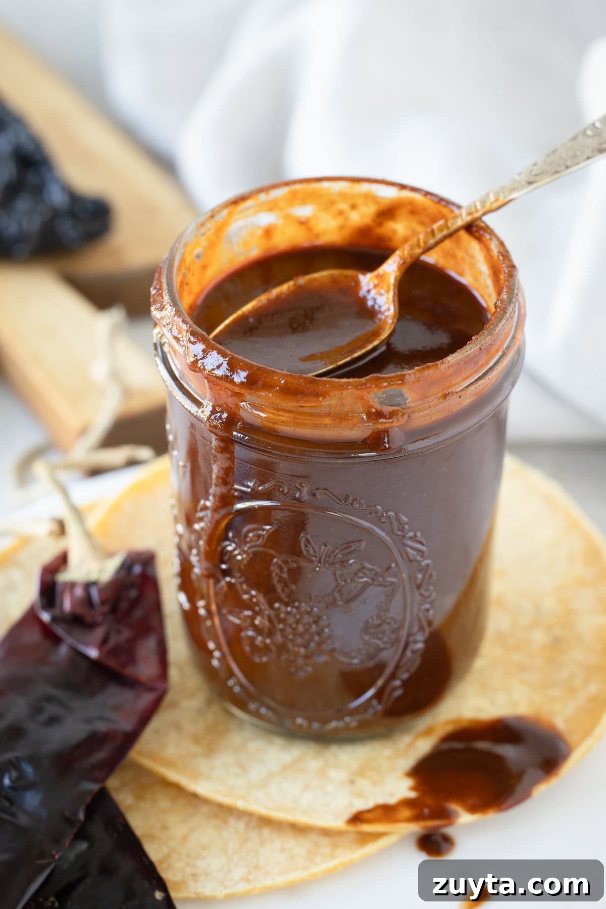 A glass jar of deep brick red enchilada sauce with a messy spoon, and dried chiles and tortillas.